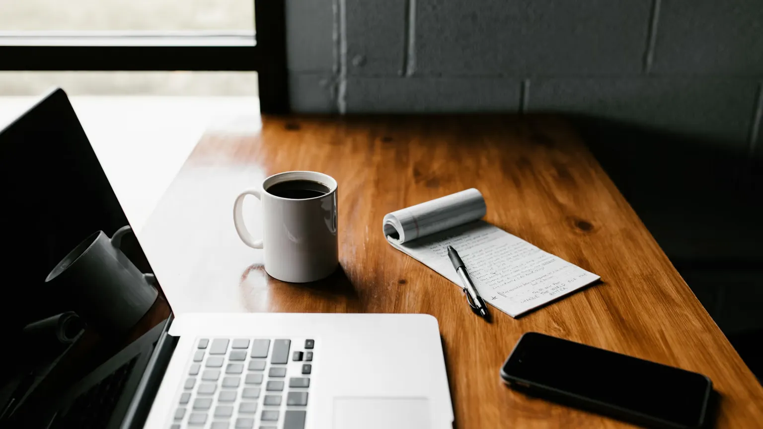 A laptop, a mug of black coffee, a phone and a notepad of handwritten notes on a wooden desk